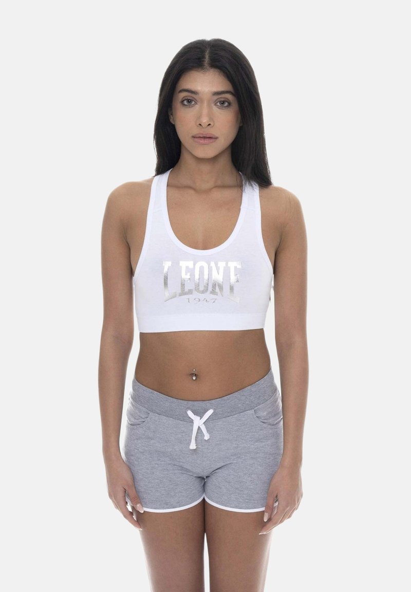 Femme aux longs cheveux foncés portant un soutien-gorge de sport blanc avec le texte "LEONE 1947" et un short gris avec cordon, debout devant un fond uni.
