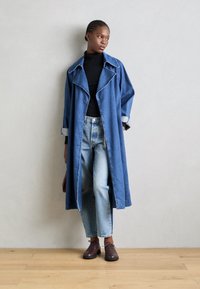 Cappotto lungo in denim con colletto, di colore blu, con bottoni e maniche arrotolate, abbinato a jeans azzurri e scarpe marroni.