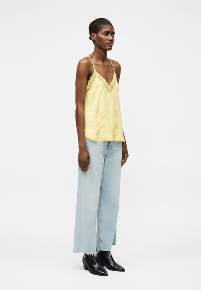 Γυναίκα που φορά κίτρινη κορσέ με δαντέλα, ανοιχτό μπλε τζιν με φαρδιά παντελόνια και ακατέργαστα τελειώματα, καθώς και μαύρα ankle boots με μυτερή μύτη, που στέκεται πλαγίως.
