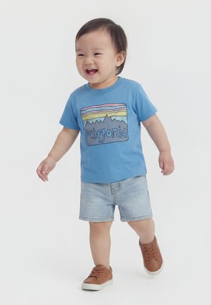 Väikelaps kõnnib ja naeratab, kandes sinist Patagonia t-särki, heledaid teksapükse ja pruune tenniseid, taustaks on ühevärviline valge taust.