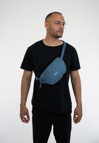 Zöld fanny pack cipzárral és állítható pánttal. Simább szövet, ívelt forma, kis logó folt elöl. Keresztben hordva.
