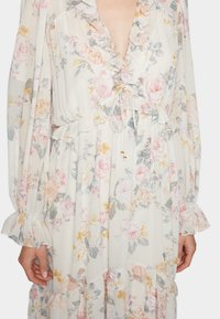 Robe florale en tissu léger, avec un col et des poignets volants, une taille à cordon et un ourlet à plusieurs niveaux, ornée de motifs floraux roses, jaunes et verts.