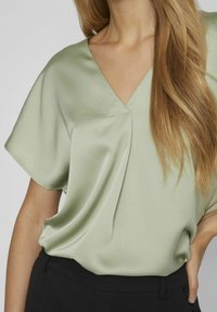 Blusa di raso verde chiaro con scollo a V, maniche corte e morbidi drappeggi, abbinata a pantaloni neri. Texture liscia, lucentezza sottile.
