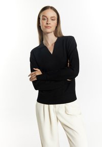 Black knit sweater with a V-neck and collar, featuring long sleeves and a fitted waistband, paired with cream pleated trousers.