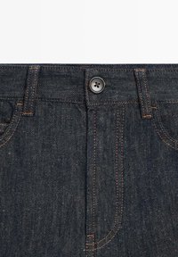 Granatowa talia spodni dżinsowych z miedzianym przeszyciem, zapięciem na guzik, szlufkami na pasek i widocznymi przednimi kieszeniami.