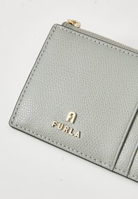 Hellgraue Lederbörse mit einer strukturierten Oberfläche, ausgestattet mit einem goldenen "FURLA"-Logo und einem Reißverschlussfach. Kompakte und rechteckige Form.