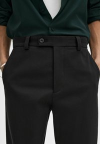 Pantaloni neri realizzati in tessuto a trama, con chiusura a bottone, tasche laterali e superficie liscia. Abbinati a una camicia verde con bottoni.