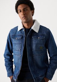 Jeansjacke in Dunkelblau mit einem weißen Kunstfellkragen. Hat zwei Brusttaschen und einen Reißverschluss vorne. Strukturierter Stoff.