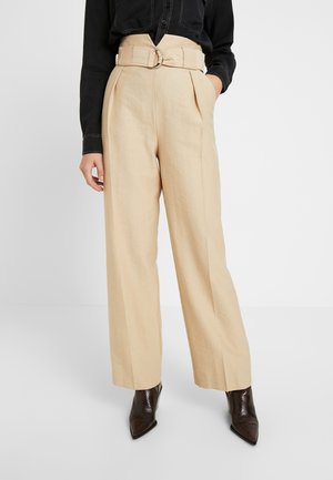 Personne portant un pantalon large taille haute beige avec une ceinture assortie et des bottines noires à talons pointus, main dans la poche, sur fond blanc.