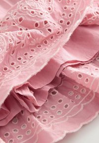 Tissu rose avec une broderie complexe et des motifs découpés, bordures festonnées, texture douce avec une apparence légèrement froissée.