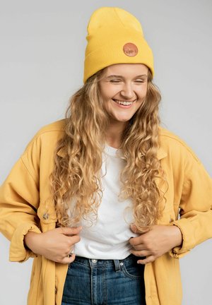 Żółta wełniana czapka z okrągłą skórzaną naszywką, połączona z pasującą żółtą kurtką i białą koszulką, a także ciemnoniebieskimi dżinsami. Długie, kręcone włosy.