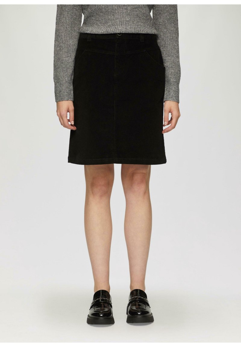 Femme portant une jupe en velours côtelé noire longueur genou, un pull en tricot gris et des mocassins en cuir verni noir, debout devant un fond uni.