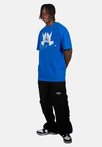 Kék pamut póló, amelyen két fehér madár grafikája és alatta a "YOUTH" felirat található. Fekete cargo nadrággal és sportcipővel párosítva.