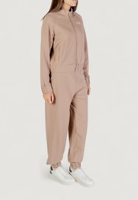 Chándal beige con cuello alto y cierre en la parte delantera. La parte superior presenta un logo en el pecho; los pantalones tienen un corte relajado con puños elásticos. Combinado con zapatillas blancas.