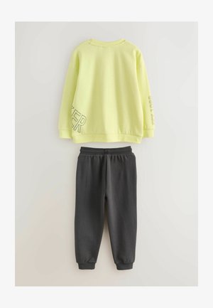 Svetlo rumena dukserica s okruglim izrezom, sa ispisanim tekstom na rukavu i bočnoj strani; u kombinaciji s tamnosivim duksericama s elastičnim strukom i suženim gležnjevima.