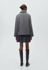 Manteau en laine gris mélangé avec un grand col, vu de dos. Associé à une robe à carreaux foncés et à de hautes bottes en cuir noires.