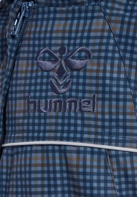 Blåt ternet stof med brune accenter; har en lynlås, hvid piping og det broderede "hummel" logo med et grafisk design ovenfor.