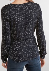 Blouse noire à manches longues avec des pois blancs, dotée d'un col rond, de détails froncés à la taille et de poignets boutonnés.
