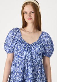 Μπλε φλοράλ φόρεμα με φουσκωτά μανίκια, συγκεντρωμένο ντεκολτέ και φούστα με στιβαρές πτυχώσεις. Έχει ελαφρύ μπλε σχέδιο σε πιο σκούρο μπλε φόντο.