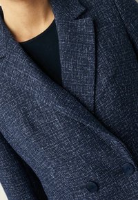 Chaqueta texturizada azul marino con patrón de sarga, solapas con muesca y dos botones negros. Lleva puesta sobre un top negro, con énfasis en el detalle de la tela.