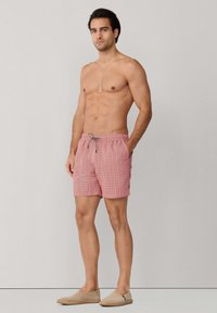 Homme torse nu portant un short de bain rouge à motifs avec un cordon de serrage, des espadrilles beiges, debout devant un fond gris clair uni.