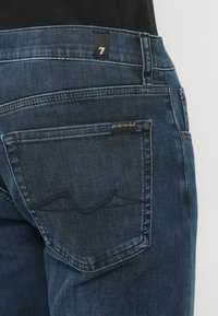 Vaqueros de mezclilla azul oscuro con un bolsillo trasero que muestra un diseño de ola cosido y una etiqueta que dice "for all man kind". Primer plano de la cinturilla.