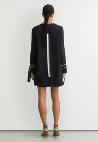 Tumši zila kleita ar garām piedurknēm, taisnu siluetu, baltām vertikālām svītrām muguras pusē un sasienamiem akcentiem piedurkņu galiem.