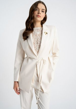 Kobieta ubrana w kremowy, dopasowany garnitur z koronkową bluzką, paskiem w talii i złotą broszką na lewym klapie marynarki na jednolitym tle.