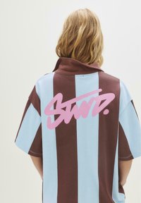 Person med skulderlangt lyst hår iført en blå og brun stribet skjorte med stor pink tekst på ryggen.