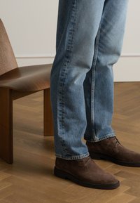 Bottes en suède marron à bout rond, à lacets, portées avec un jean bleu clair, se tenant à côté d'une chaise en bois sur un sol en parquet.