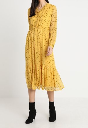 Kvinde iført gul midi-kjole med lange ærmer, sort plettet mønster og flæset kant, kombineret med sorte ankelstøvler med hæl.