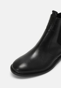 Černé kožené Chelsea boty s kulatou špičkou, elastickou boční klopou a poutkem na bílem pozadí.