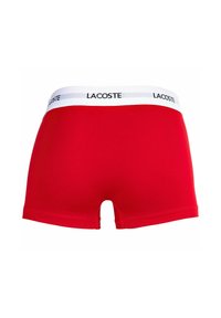Červené bavlnené boxerky s bielym pásom a čiernym brandingom "LACOSTE". Dizajn obsahuje krátke nohy a priliehavý strih.