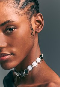 Strieborný náhrdelník s čiernymi a bielymi kruhovými diskami, ktoré sa striedajú a sú spojené kovovými krúžkami, ležiaci na hladkej pokožke s viditeľnými vrkôčikmi.