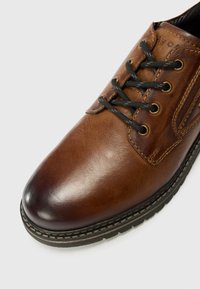 Brown leather shoe with a rounded toe, black laces, stitching details, and a textured finish. Padded side area enhances comfort.