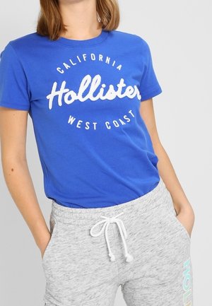 Osoba nosząca niebieską koszulkę "Hollister California West Coast" oraz jasnoszare spodnie dresowe ze sznurkiem, z rękami w kieszeniach.