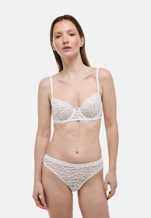 Femme portant un soutien-gorge en dentelle blanche et une culotte assortie, debout devant un fond clair uni.