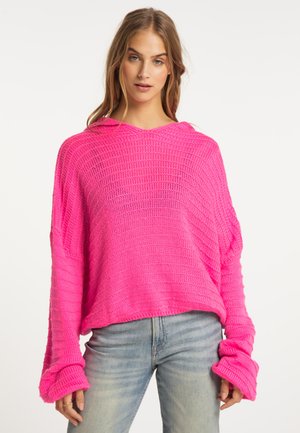 Frau mit langen Haaren, die einen hellrosa locker gestrickten Pullover und hellblaue Jeans trägt, vor einem schlichten weißen Hintergrund stehend.