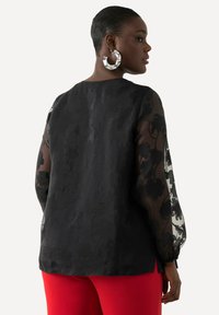 Schwarze Bluse mit durchsichtigen, gemusterten Ärmeln, strukturiertem Stoff am Körper und Rundhalsausschnitt, kombiniert mit roten Hosen.