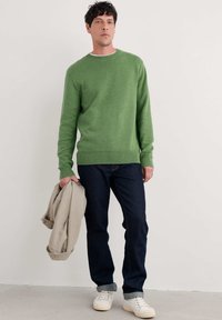 Suéter de punto verde con cuello redondo, combinado con jeans de mezclilla oscura y zapatillas blancas, sosteniendo una chaqueta de color beige claro.