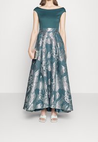 Robe de soirée teal avec décolleté bardot, jupe texturée à motifs argentés, associée à des sandales vert menthe et une pochette argentée.