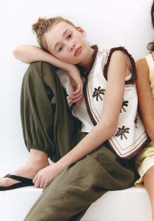 Giovane donna con chignon è seduta a piedi nudi con pantaloni verdi e gilet bianco con ricamo a palma, appoggiando la testa sul ginocchio su sfondo bianco.