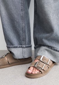 Sandales marron avec deux larges sangles, boucles en argent et clous en argent. Portées avec un pantalon en denim gris retroussé, mettant en avant un look décontracté.