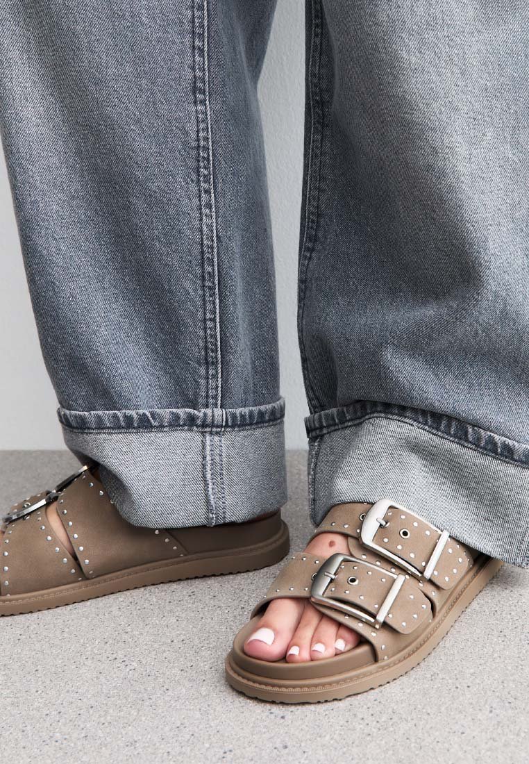 Sandales marron avec deux larges sangles, boucles en argent et clous en argent. Portées avec un pantalon en denim gris retroussé, mettant en avant un look décontracté.