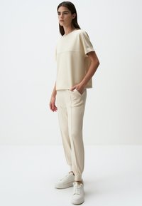Beige katoenen blend top met korte mouwen, gecombineerd met bijpassende tapse broek met zijzakken. Model draagt witte sneakers.