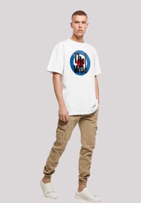 Weißes Baumwoll-T-Shirt mit einem runden Grafikdesign in Blau und Rot, kombiniert mit beigen Cargo-Hosen und weißen Sneakers.