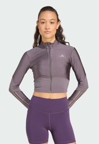 Lila kort zip-up jacka gjord av ett slätt tyg, med långa ärmar som har tre vertikala ränder på varje arm, tillsammans med högmidjade lila shorts.