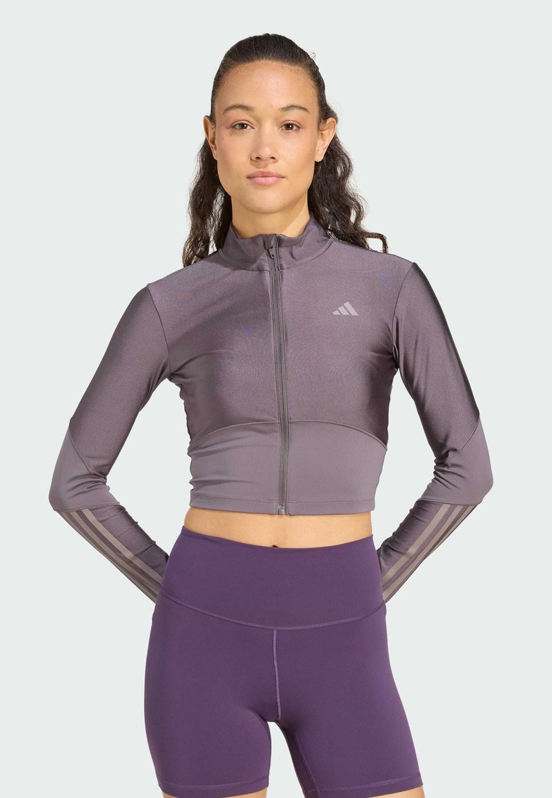 Lila kort zip-up jacka gjord av ett slätt tyg, med långa ärmar som har tre vertikala ränder på varje arm, tillsammans med högmidjade lila shorts.