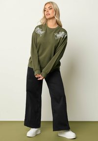 Felpa verde oliva con dettagli ricamati floreali sulle spalle, abbinata a pantaloni neri a vita alta e sneakers bianche.