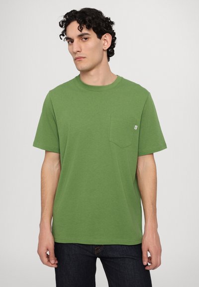 Green cotton t-shirt with short sleeves, crew neckline, and chest pocket. Smooth texture, straight fit, worn with dark denim jeans.
